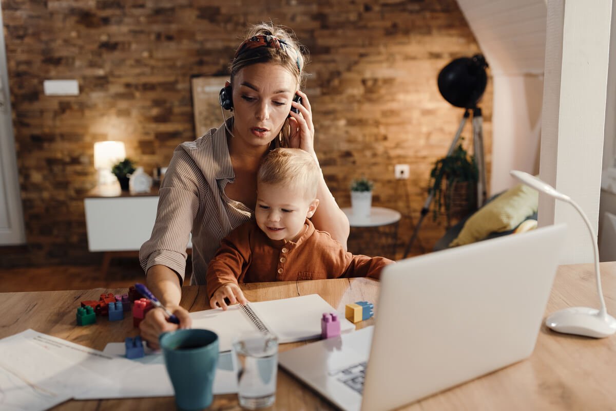 Une mère de famille gérant son ordinateur de travail à la maison tout en s'occupant de son jeune enfant.